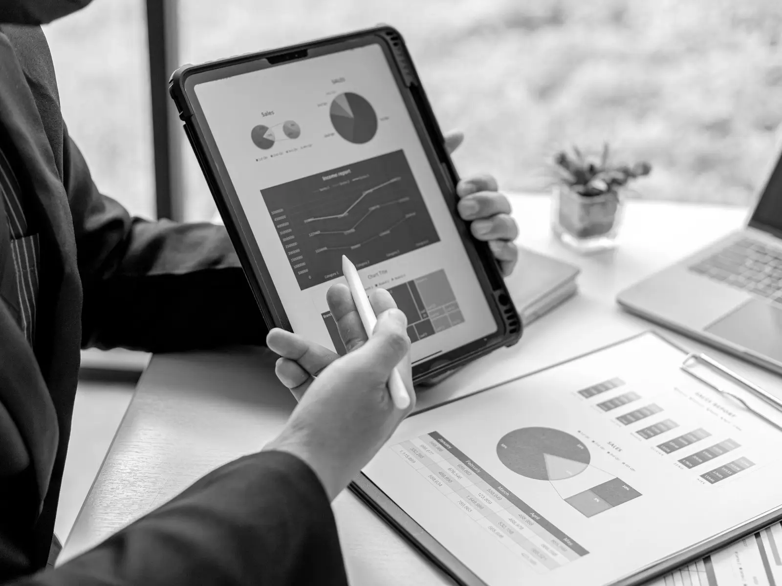 Pessoa analisando gráficos de vendas num tablet, com relatórios impressos sobre a mesa